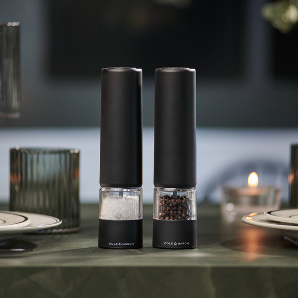Two Brixton Electric Mill Matt Black GS grinders by Cole & Mason, featuring convenient one-hand operation and a modern design, sit side by side on a green table, with plates, glassware, and a lit candle softly blurred in the background.