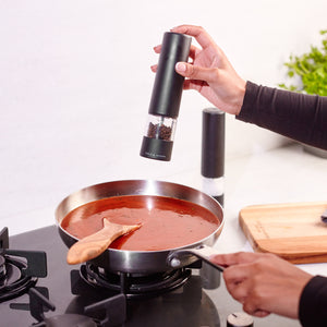 A person seasons a pan of red sauce on the stovetop using the Cole & Mason Brixton Electric Mill Matt Black GS. A wooden spoon rests in the sauce, while a cutting board and the pepper mill are on the nearby counter.
