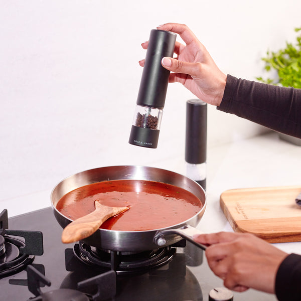A person seasons a pan of red sauce on the stovetop using the Cole & Mason Brixton Electric Mill Matt Black GS. A wooden spoon rests in the sauce, while a cutting board and the pepper mill are on the nearby counter.