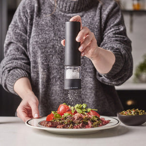 Wearing a gray sweater, a person grinds salt onto a plate of steak and vegetables with the Cole & Mason Brixton Electric Mill Matt Black GS. The plate rests on a white countertop next to a small bowl of garnish.