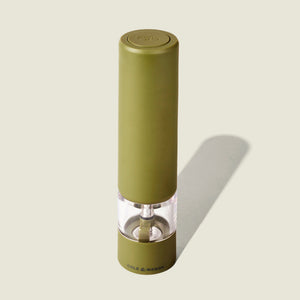 The Cole & Mason Brixton Electric Mill Olive Green GS stands upright on a light beige background, casting a shadow to the right. The green mill features a transparent section that reveals the ceramic grinder and grinding mechanism, and it is designed for convenient one-handed use.