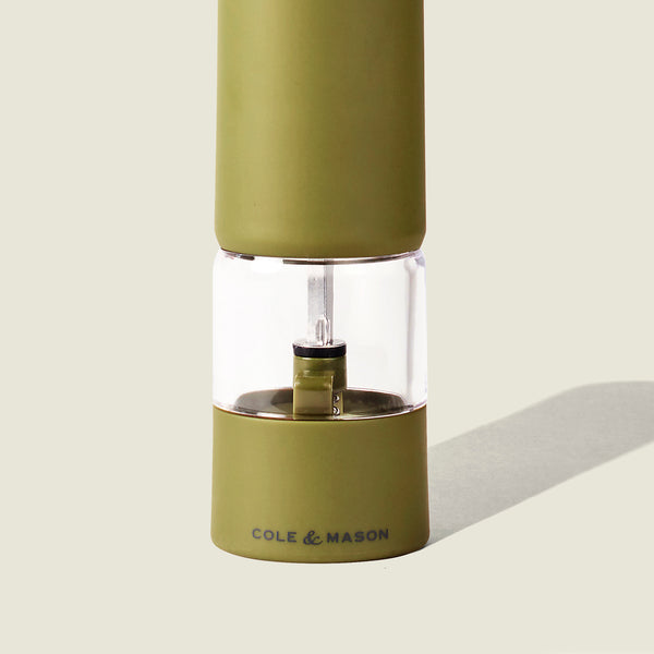 A Cole & Mason Brixton Electric Mill Olive Green GS pepper grinder, featuring a transparent middle section that reveals the ceramic grinder and internal mechanism, designed for effortless one-handed use and casting a shadow on a light background.