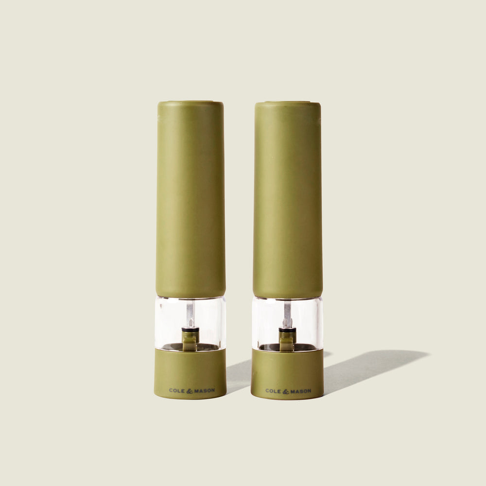 Two olive green Cole & Mason Brixton Electric Mills with clear midsections stand upright side by side on a neutral background, casting light shadows to the right. Designed for convenient one-handed use, these mills feature a durable ceramic grinder for effortless seasoning.