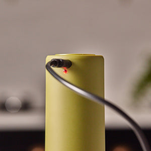 A close-up of the Cole & Mason Brixton Electric Mill Olive Green GS, a cylindrical electronic device designed for one-handed use, with a black cable plugged in and a small red indicator light turned on. The electric salt and pepper mill features a durable ceramic grinder for efficient seasoning.