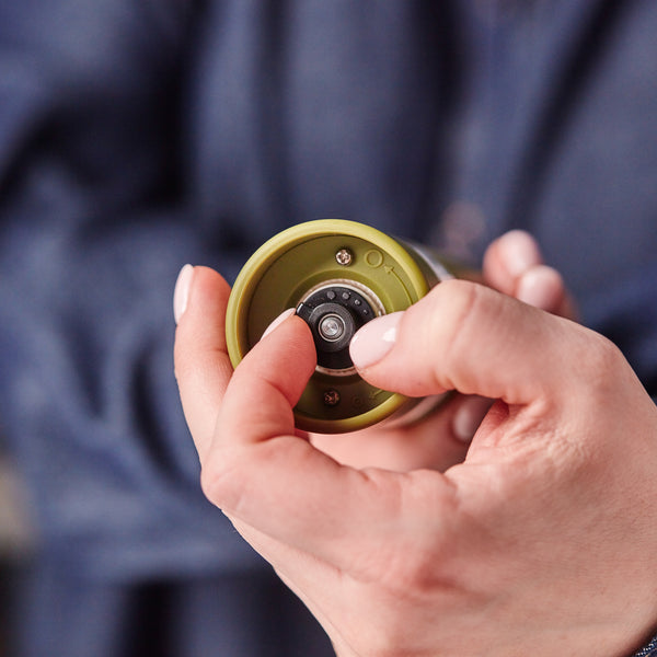 A person wearing a blue long-sleeve shirt adjusts the ceramic grinder setting on the bottom of a Cole & Mason Brixton Electric Mill Olive Green GS, using their thumb and forefinger.