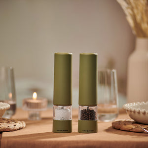 Two Brixton Electric Mill Olive Green GS salt and pepper mills by Cole & Mason, featuring durable ceramic grinders, sit on a beige tablecloth surrounded by glassware, plates, and a lit tealight candle in a cozy, softly lit dining setting. Designed for effortless one-handed use, these stylish electric mills add both convenience and elegance to your table.