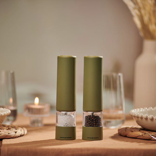 Two Brixton Electric Mill Olive Green GS salt and pepper mills by Cole & Mason, featuring durable ceramic grinders, sit on a beige tablecloth surrounded by glassware, plates, and a lit tealight candle in a cozy, softly lit dining setting. Designed for effortless one-handed use, these stylish electric mills add both convenience and elegance to your table.