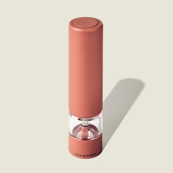 The Cole & Mason Brixton Electric Mill Terracotta GS stands upright on a light beige background, its pink body and transparent middle section showcasing the ceramic grinding system inside. This sleek electric pepper grinder offers convenient USB-C charging.