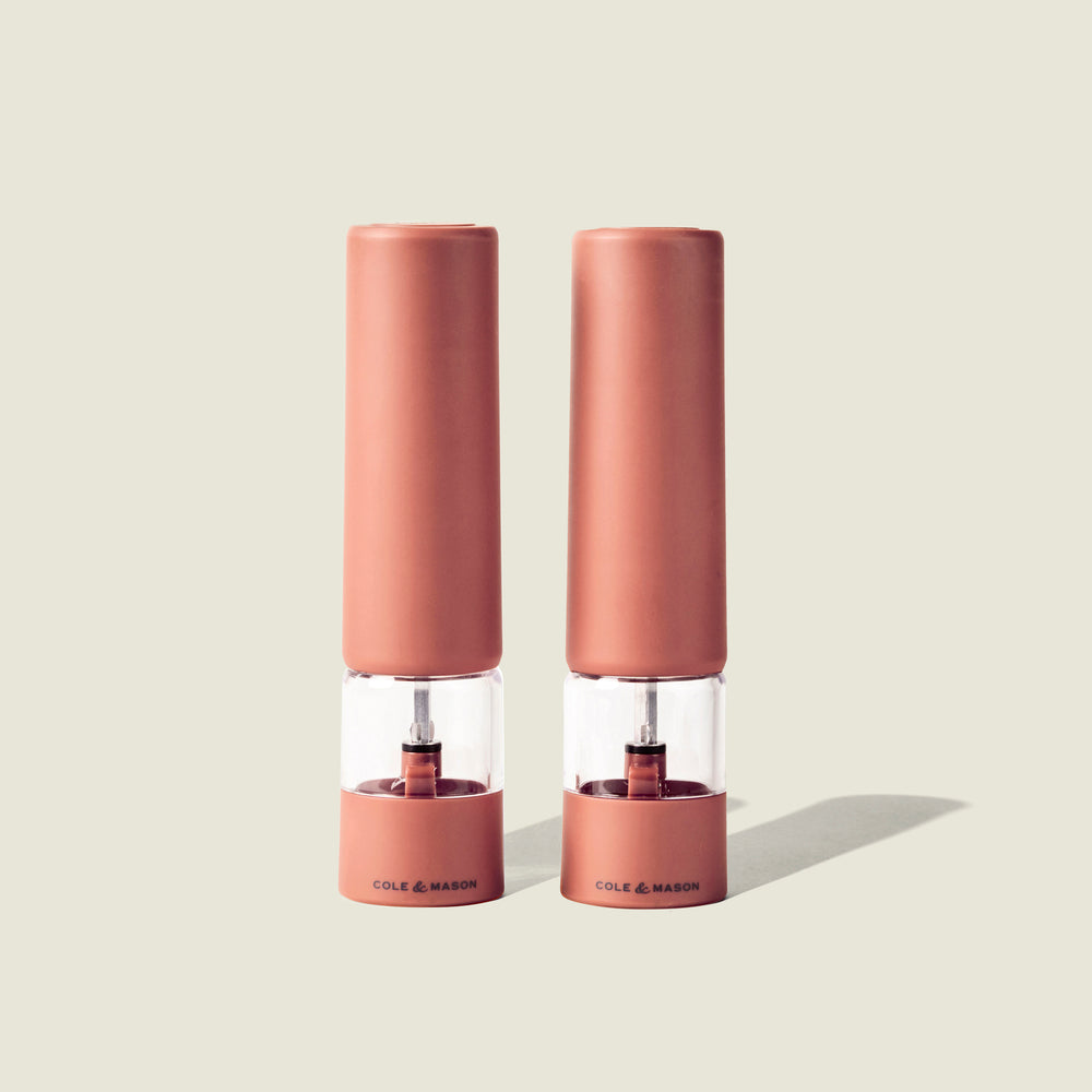 Two identical Cole & Mason Brixton Electric Mill Terracotta GS grinders stand side by side on a light beige background, casting soft shadows to the right. Featuring a durable ceramic grinding system and convenient USB-C charging, these mills combine modern functionality with elegant design.