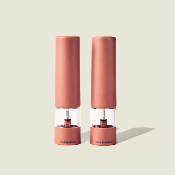 Two identical Cole & Mason Brixton Electric Mill Terracotta GS grinders stand side by side on a light beige background, casting soft shadows to the right. Featuring a durable ceramic grinding system and convenient USB-C charging, these mills combine modern functionality with elegant design.