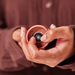 A person with manicured nails holds the Cole & Mason Brixton Electric Mill Terracotta GS—a small, round, terracotta-colored device featuring a central camera-like lens and subtle hints of sleek technology that reveal its rechargeable design with USB-C compatibility. Both the background and the person’s clothing are brown, creating a warm, cohesive palette.