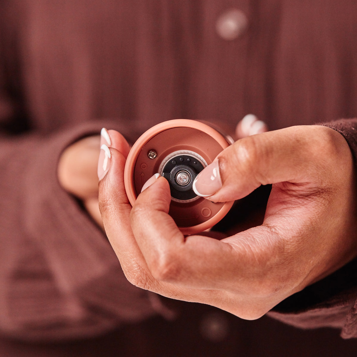 A person with manicured nails holds the Cole & Mason Brixton Electric Mill Terracotta GS—a small, round, terracotta-colored device featuring a central camera-like lens and subtle hints of sleek technology that reveal its rechargeable design with USB-C compatibility. Both the background and the person’s clothing are brown, creating a warm, cohesive palette.