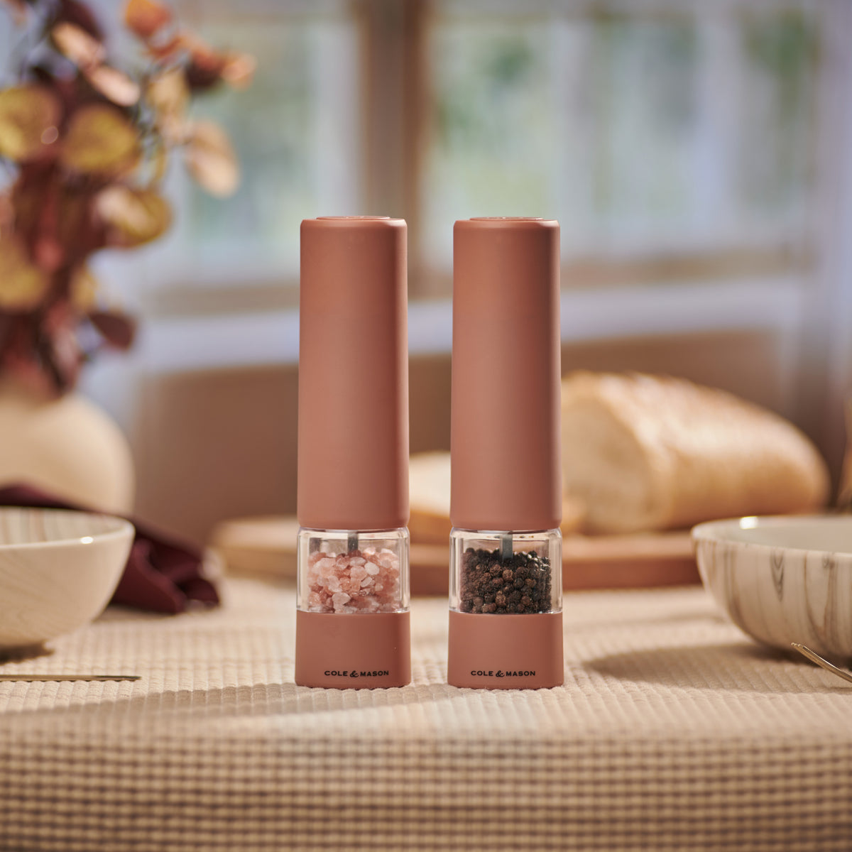 Two Cole & Mason Brixton Electric Mill Terracotta GS grinders, filled with pink Himalayan salt and black peppercorns, sit on a beige tablecloth. Their ceramic grinding system delivers both style and functionality to any setting. In the background, a loaf of bread and bowls are visible as sunlight streams through a window.