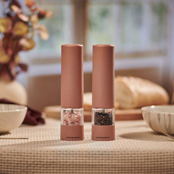 Two Cole & Mason Brixton Electric Mill Terracotta GS grinders, filled with pink Himalayan salt and black peppercorns, sit on a beige tablecloth. Their ceramic grinding system delivers both style and functionality to any setting. In the background, a loaf of bread and bowls are visible as sunlight streams through a window.