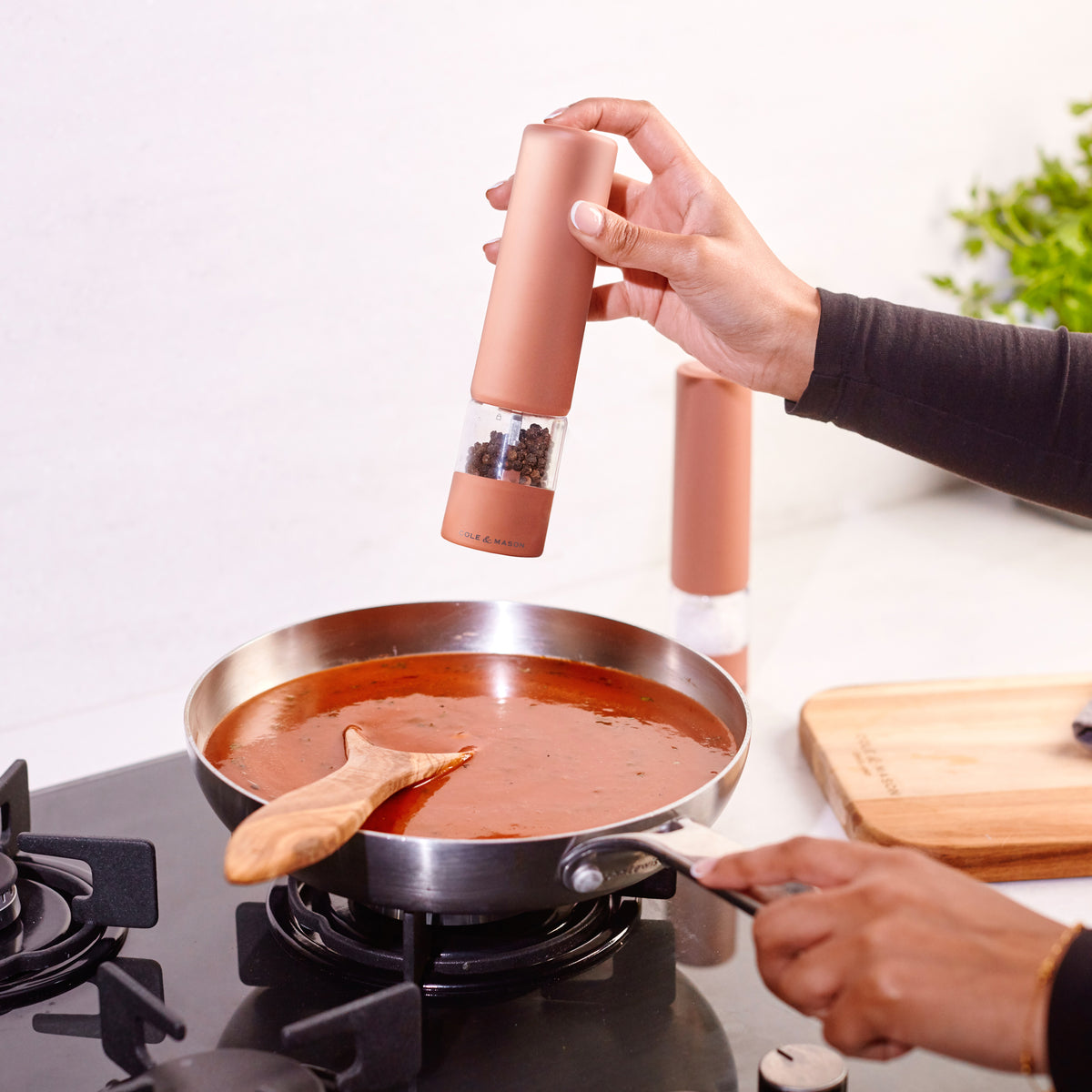 A person sprinkles pepper into a pan of tomato sauce on a stove, stirring with a wooden spoon. In the background, a cutting board and the Cole & Mason Brixton Electric Mill Terracotta GS—featuring a ceramic grinding system and convenient USB-C charging port—are visible.