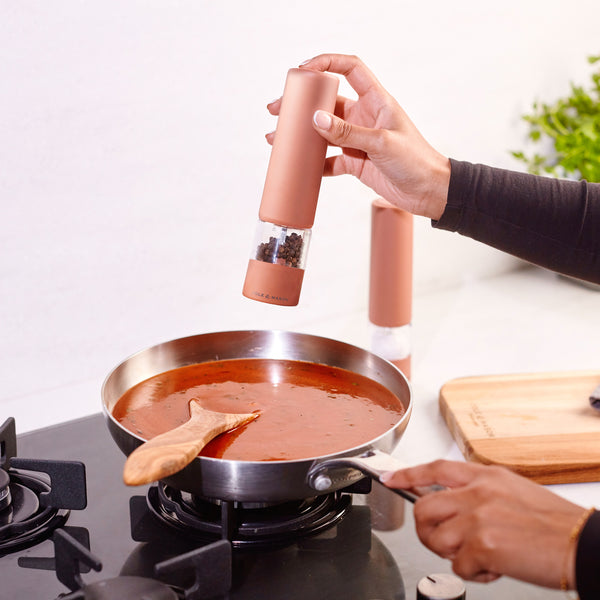 A person sprinkles pepper into a pan of tomato sauce on a stove, stirring with a wooden spoon. In the background, a cutting board and the Cole & Mason Brixton Electric Mill Terracotta GS—featuring a ceramic grinding system and convenient USB-C charging port—are visible.