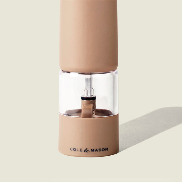 A tan Cole & Mason Brixton Electric Mill Stone GS pepper grinder with a clear middle section, featuring an adjustable grind and a durable ceramic grinding core, shown against a light beige background.