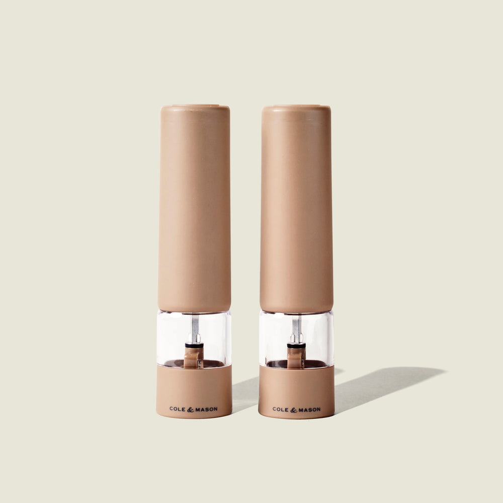 Two beige Cole & Mason Brixton Electric Mill Stone GS grinders with transparent midsections and the brand name printed at their bases, featuring adjustable grind settings and durable ceramic grinding cores, stand side by side on a light neutral background.