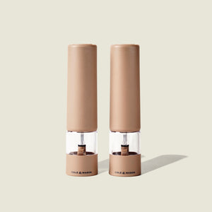 Two beige Cole & Mason Brixton Electric Mill Stone GS grinders with transparent midsections and the brand name printed at their bases, featuring adjustable grind settings and durable ceramic grinding cores, stand side by side on a light neutral background.
