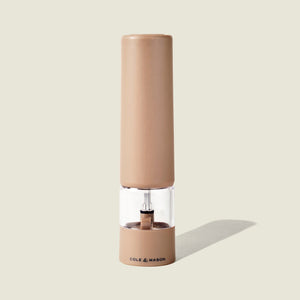 A tan-colored Brixton Electric Mill Stone GS pepper grinder by Cole & Mason stands upright on a light beige background, casting a shadow to the right. It features a transparent middle section, an adjustable grind setting, and a durable ceramic grinding core. The Cole & Mason brand is printed on the bottom front.