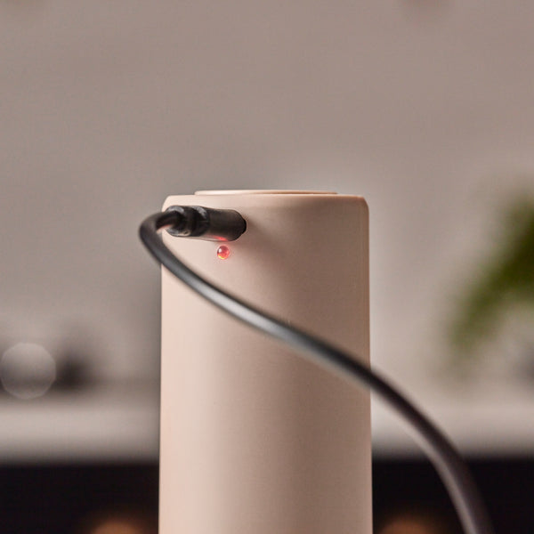 A close-up of the Cole & Mason Brixton Electric Mill Stone GS shows its beige finish and ceramic grinding core, with a black charging cable plugged in and a small red LED indicator light illuminated near the connector.