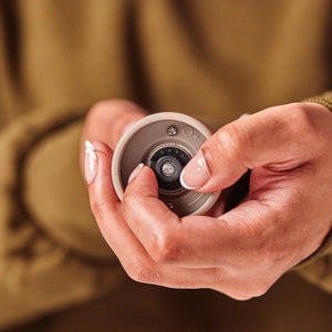 A person with manicured nails holds a small round camera lens device, focusing closely on the lens. The background is blurred and the person is wearing a brown long-sleeved top, echoing the refined craftsmanship of the Cole & Mason Brixton Electric Mill Stone GS, which features an adjustable grind and a ceramic grinding core.