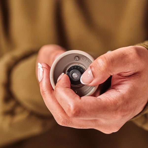 A person with manicured nails holds a small round camera lens device, focusing closely on the lens. The background is blurred and the person is wearing a brown long-sleeved top, echoing the refined craftsmanship of the Cole & Mason Brixton Electric Mill Stone GS, which features an adjustable grind and a ceramic grinding core.