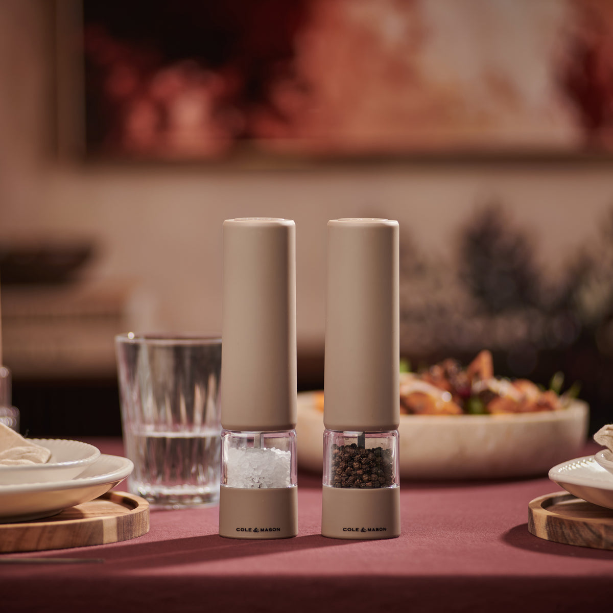 Two beige Cole & Mason Brixton Electric Mill Stone GS salt and pepper grinders with ceramic grinding cores sit on a dining table set with plates, glasses, and food, with a blurred background.