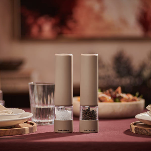 Two beige Cole & Mason Brixton Electric Mill Stone GS salt and pepper grinders with ceramic grinding cores sit on a dining table set with plates, glasses, and food, with a blurred background.