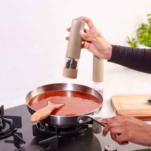 A person seasons tomato sauce in a pan using the Cole & Mason Brixton Electric Mill Stone GS, an electric pepper grinder with adjustable grind settings, while holding the pan handle; a wooden spoon rests in the sauce. A cutting board and fresh herbs are visible on the kitchen counter in the background.