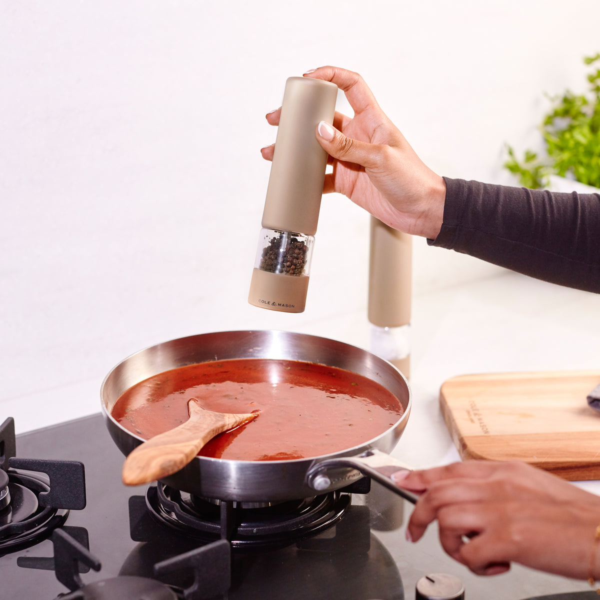 A person seasons tomato sauce in a pan using the Cole & Mason Brixton Electric Mill Stone GS, an electric pepper grinder with adjustable grind settings, while holding the pan handle; a wooden spoon rests in the sauce. A cutting board and fresh herbs are visible on the kitchen counter in the background.