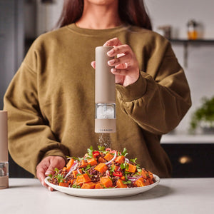 A person in a brown sweater is using the Cole & Mason Brixton Electric Mill Stone GS, which features an adjustable grind, to season a colorful salad on a white plate in a kitchen.