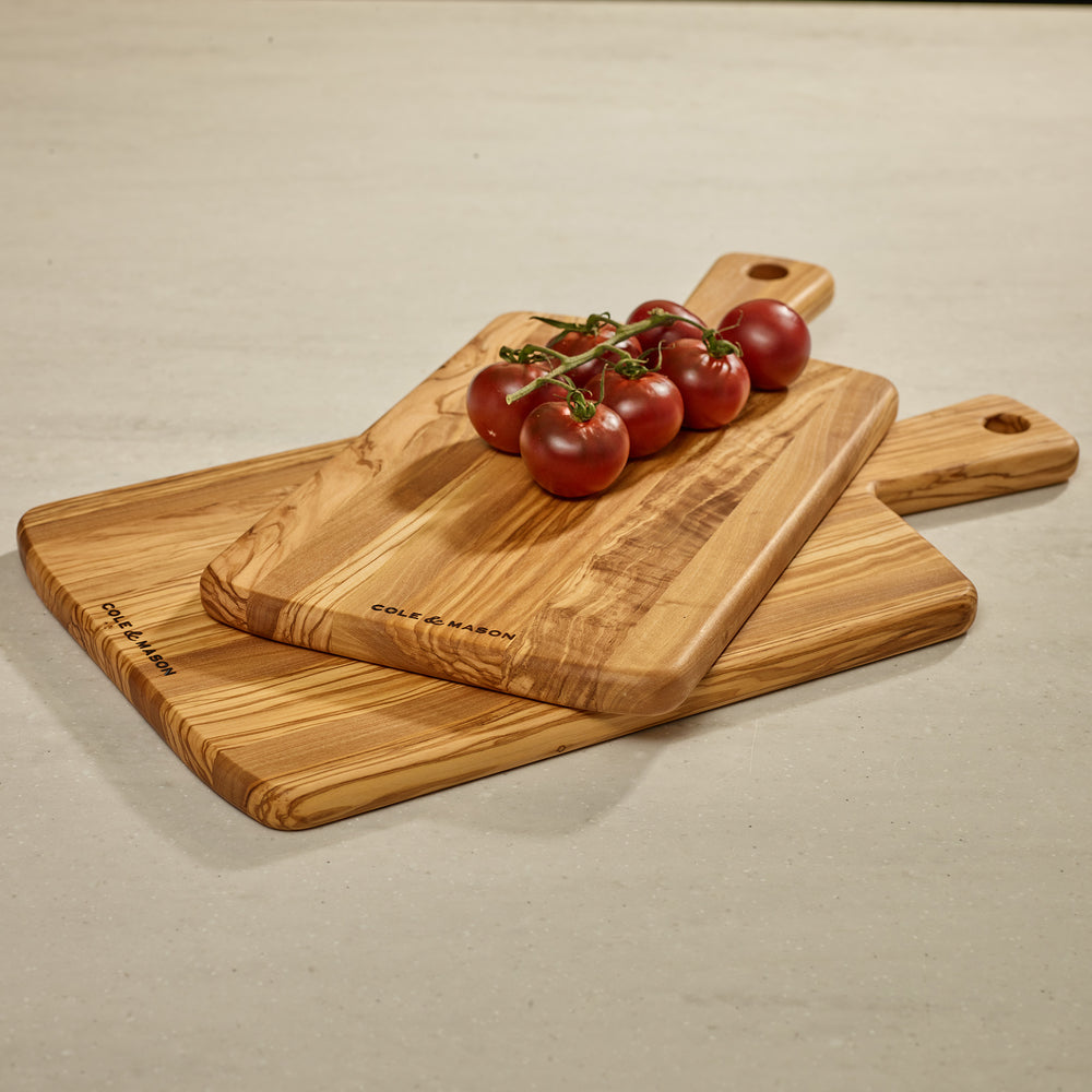 The Cole & Mason Portobello Olive Wood Serving Board, with a handle and hanging hole, showcases an elegant natural grain—ideal as a stylish antipasti board and shown upright on a light background.
