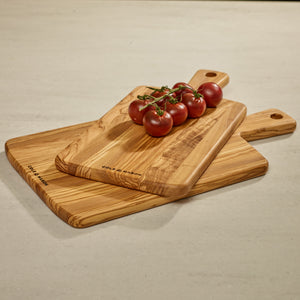 Three Cole & Mason Portobello Olive Wood Serving Boards are stacked on a light countertop, each with a handle and engraved branding. A cluster of fresh red vine tomatoes rests atop the uppermost board.
