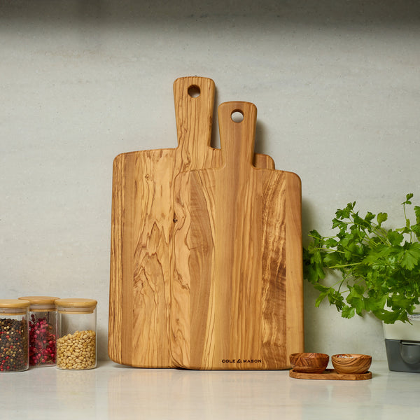 The Cole & Mason Portobello Olive Wood Serving Boards Set stands upright on a kitchen counter beside spice and bean jars, a small plant, and salt and pepper bowls, against a light gray wall.