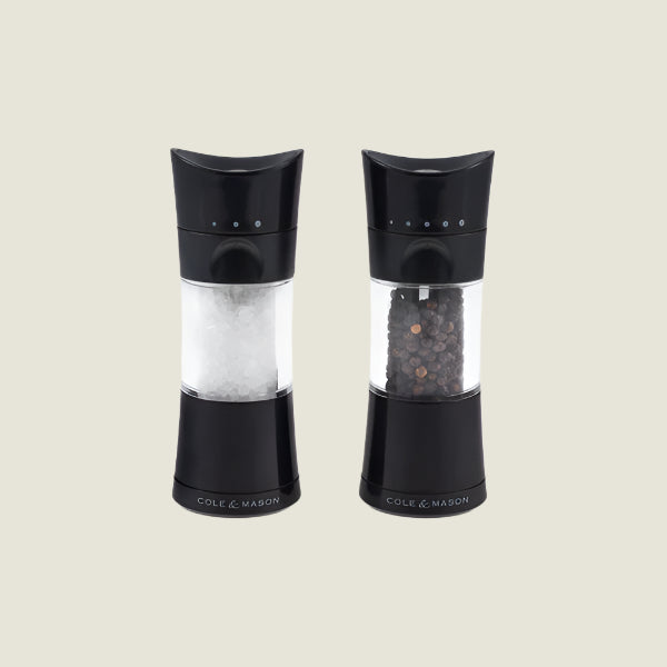 The Cole & Mason Harrogate Salt & Pepper Mill Set features stylish grinders with black accents and adjustable settings—one filled with coarse salt, the other with whole peppercorns—both upright on a plain light background.