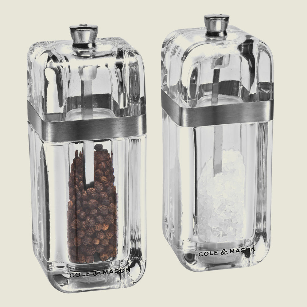 The Cole & Mason UK Kempton Salt & Pepper Mill with Refill Set 130mm features two clear rectangular mills with metal bands and knobs, filled with black peppercorns and coarse sea salt—an elegant Precision+ range gift set.