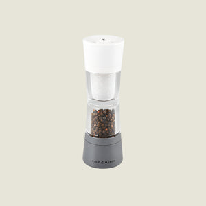 A clear and gray Cole & Mason Lincoln 2-in-1 Duo Mill spice grinder with adjustable settings, filled with peppercorns and featuring a white top, is shown against a light beige background.
