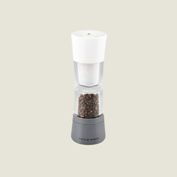 A clear and gray Cole & Mason Lincoln 2-in-1 Duo Mill spice grinder with adjustable settings, filled with peppercorns and featuring a white top, is shown against a light beige background.