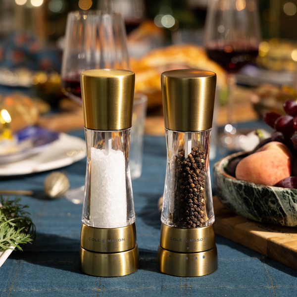 A Cole & Mason Derwent Mill Set with acrylic tray and funnel sits on a blue tablecloth, next to grapes and peaches, with blurred dining items in the background.