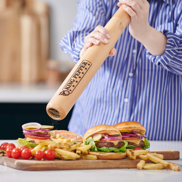 Wearing a blue striped shirt, a person seasons two burgers with fries and salad using the Cole & Mason King Pepper Baseball Bat Pepper Mill 710mm on a kitchen counter.