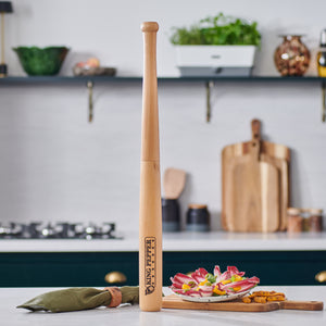 A Cole & Mason King Pepper Baseball Bat Pepper Mill 710mm stands upright on the kitchen counter, surrounded by a green pouch, appetizers, and vegetables, with shelves and utensils in the background—a perfect place for your adjustable grinder.