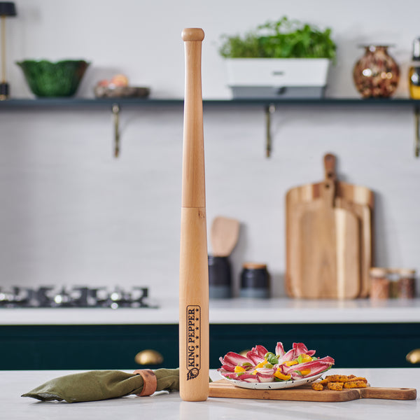 A Cole & Mason King Pepper Baseball Bat Pepper Mill 710mm stands upright on the kitchen counter, surrounded by a green pouch, appetizers, and vegetables, with shelves and utensils in the background—a perfect place for your adjustable grinder.