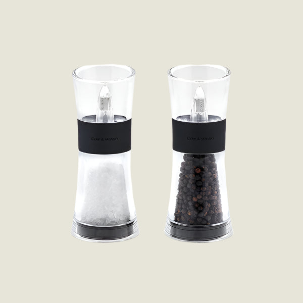 The Cole & Mason UK Flip Inverta Salt & Pepper Mill Set Black 155mm features two clear mills with black bands, one for coarse salt and one for whole black peppercorns, each with a quality grinding mechanism, shown on a light background.