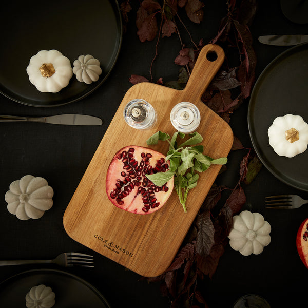 The Cole & Mason 2 Piece Barkway Board Bundle, topped with half a pomegranate, herbs, and salt and pepper shakers, rests on a dark table surrounded by white pumpkins, plates, and cutlery—a true kitchen essential.