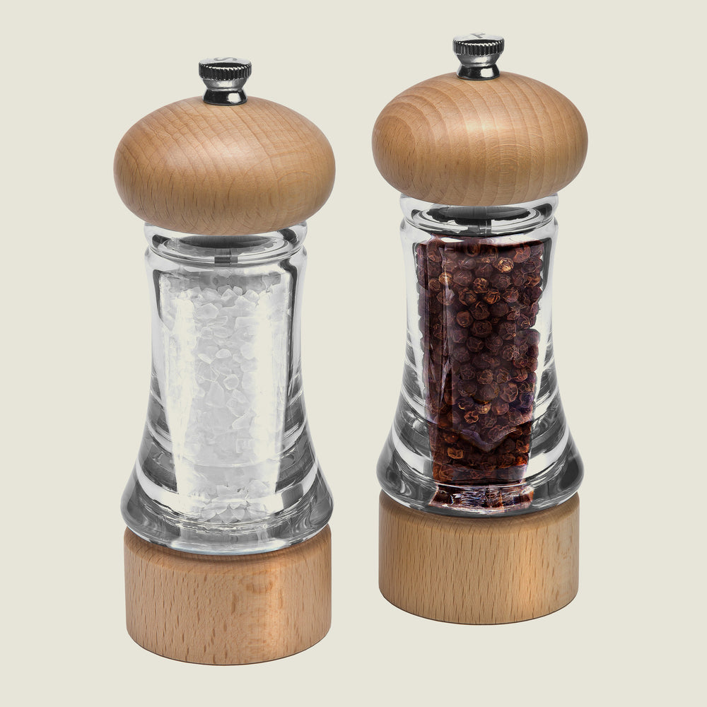 The Cole & Mason Basics Wood Salt & Pepper Mill Set features two clear mills with sturdy wood tops and bottoms, each with adjustable grind settings—one for coarse salt and one for whole black peppercorns—displayed side by side.