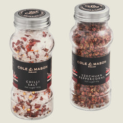 The Cole & Mason Aromatic Salt & Peppercorns Gift Set features two clear jars with silver lids, each labeled in black with red and white accents—one contains chili salt and the other Szechuan peppercorns.