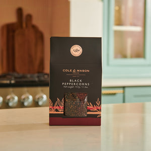 A Cole & Mason Black Peppercorns Box rests on a kitchen counter—ideal for refilling pepper mills or seasoning dishes. In the background, a stove and blue cabinets appear under gentle natural light.