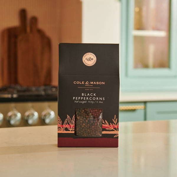 A Cole & Mason Black Peppercorns Box rests on a kitchen counter—ideal for refilling pepper mills or seasoning dishes. In the background, a stove and blue cabinets appear under gentle natural light.