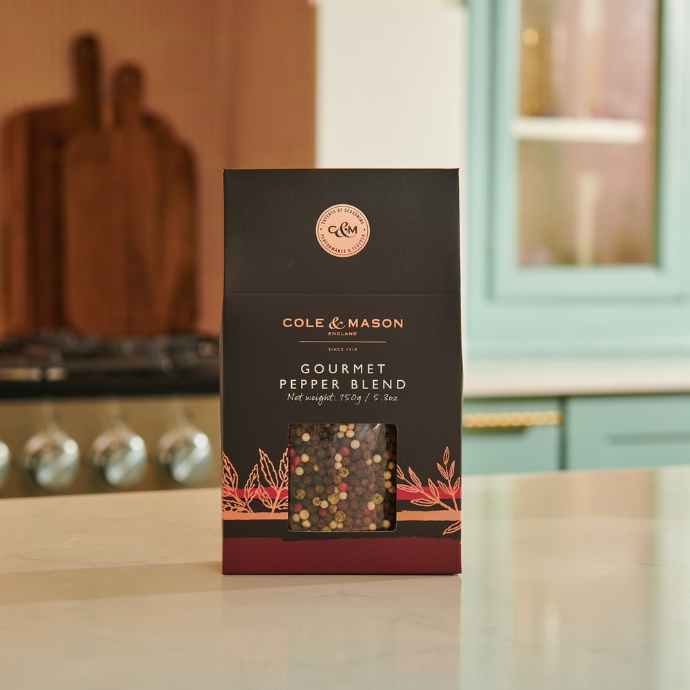 A box of Cole & Mason Gourmet Pepper Box features a clear window displaying mixed peppercorns. The mostly black packaging with brown accents is perfect for grinding fresh flavor and includes a nutrition facts label on the side.